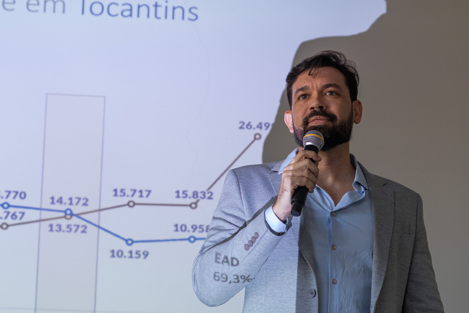 homem de barba ao microfone  com uma projeção de gráfico sobre ele e na parede ao fundo